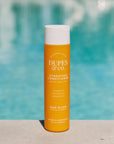 Orange bottle of DUPES & CO. conditioner with a white cap against a blurred pool background
