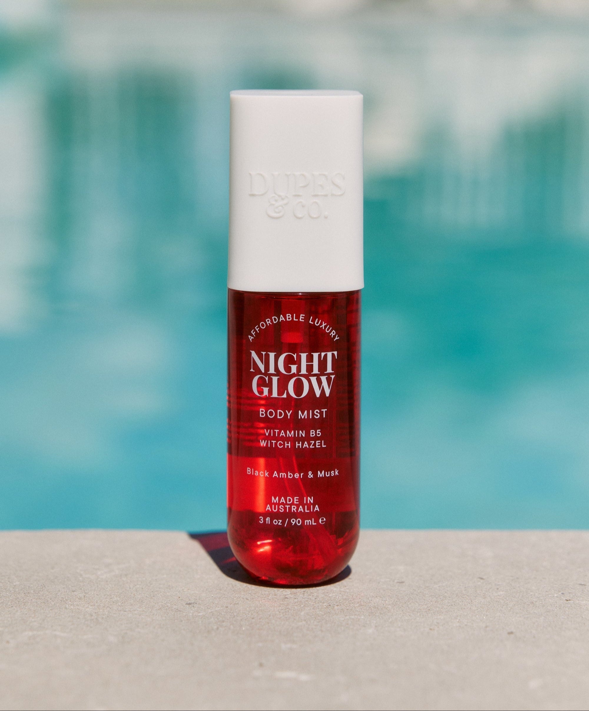 A red bottle of 'Night Glow Black Amber & Plum Body Mist' with a white cap, displayed against a poolside background.