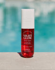 A red bottle of 'Night Glow Black Amber & Plum Body Mist' with a white cap, displayed against a poolside background.