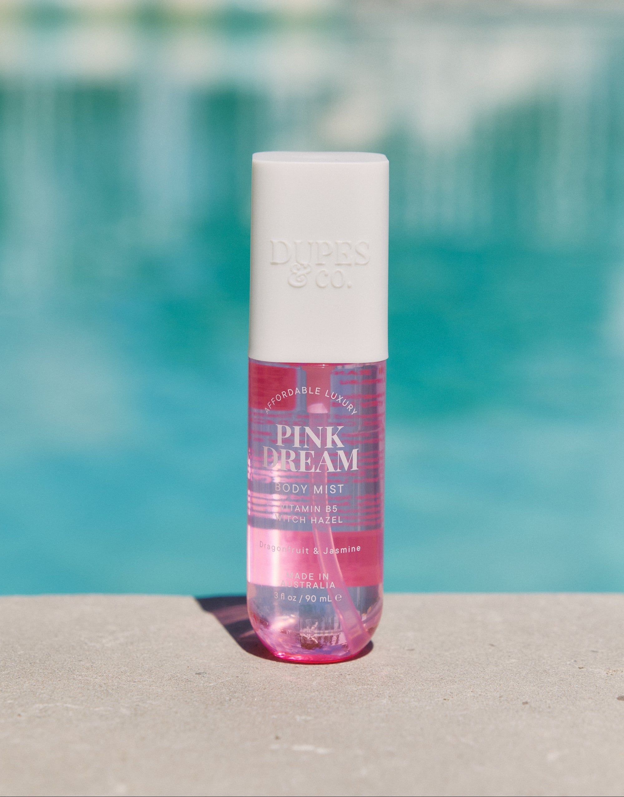 A bottle of Pink Dream body mist with pink liquid visible, placed on a surface with a pool in the background.
