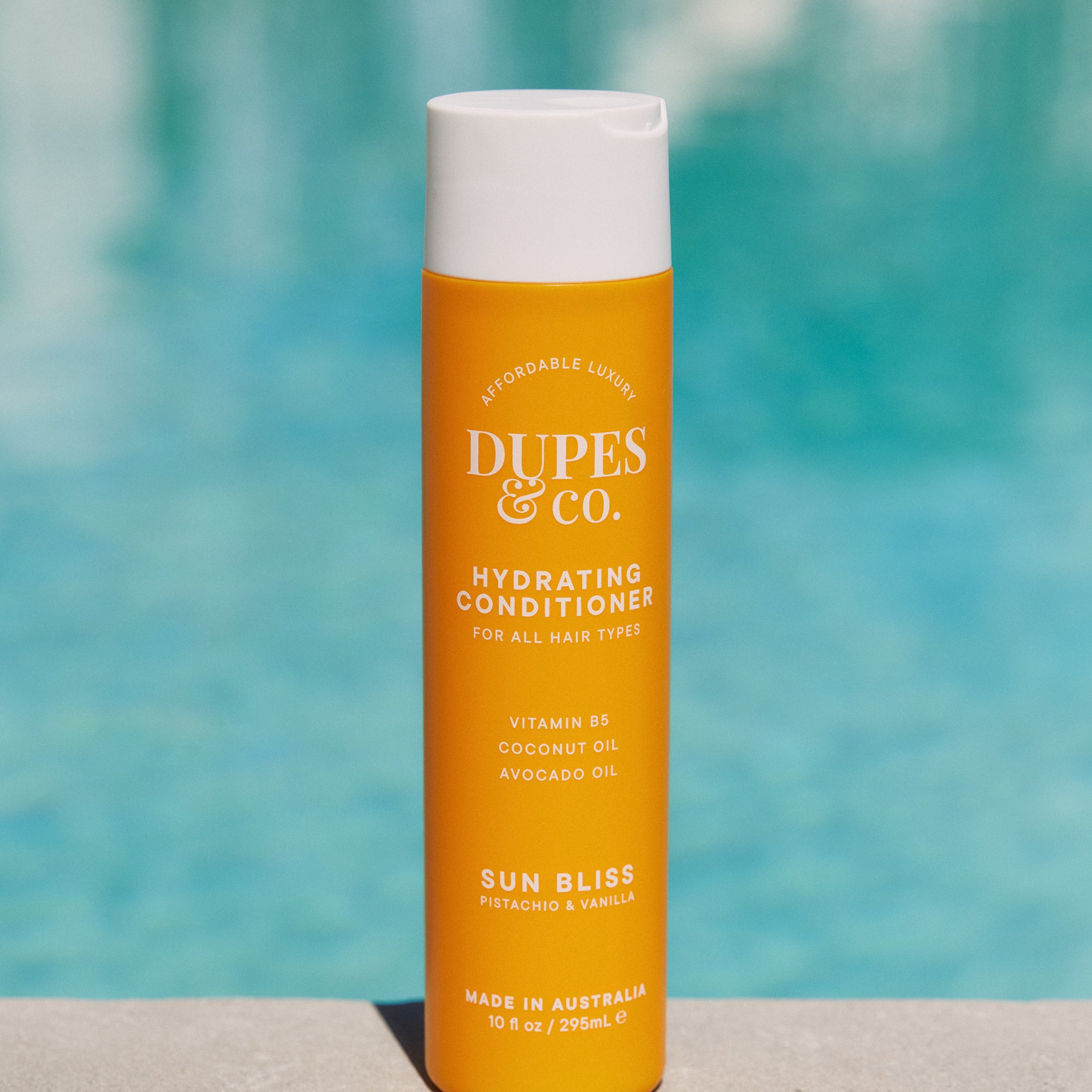 Orange bottle of DUPES & CO. conditioner with a white cap against a blurred pool background