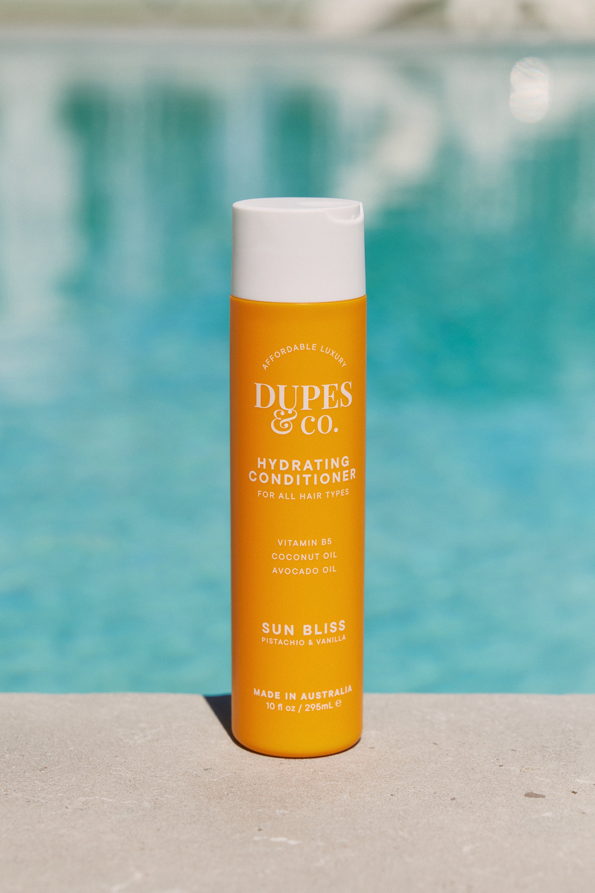 Orange bottle of DUPES & CO. conditioner with a white cap against a blurred pool background