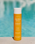 Orange shampoo bottle with white cap labeled 'Dupes & Co.' in front of a blurred pool background